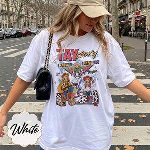 May include: A white short-sleeve t-shirt featuring a graphic print with the text TAYstory, I KNEW IT, and I KNEW YOU. The design includes three illustrated characters, a guitar, a rocket, and a block with the letters TS, all in vibrant colors.