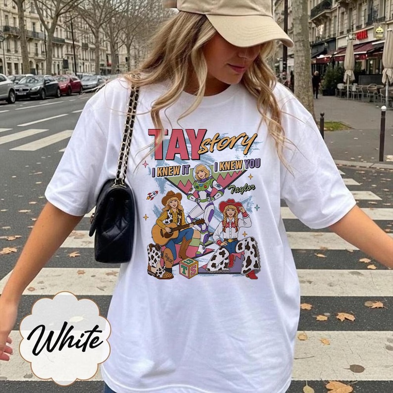 May include: A white short-sleeve t-shirt featuring a graphic print with the text TAYstory, I KNEW IT, and I KNEW YOU. The design includes three illustrated characters, a guitar, a rocket, and a block with the letters TS, all in vibrant colors.