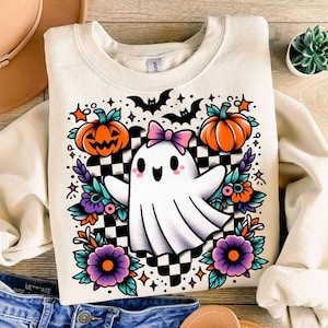 May include: Cream-colored sweatshirt featuring a Halloween-themed graphic. The design includes a cute ghost with a pink bow, pumpkins, bats, flowers, and a checkered background. The sweatshirt is folded, with a glimpse of denim jeans visible.