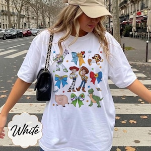 Op de afbeelding: Een wit T-shirt met korte mouwen en een kleurrijke grafische print van diverse stripfiguren, waaronder een space ranger, een cowgirl, een cowboy, een varken, een dinosaurus en een teckel, afgewisseld met decoratieve strikken en kleine sterren.