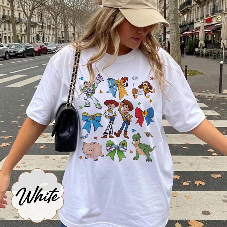 Op de afbeelding: Een wit T-shirt met korte mouwen en een kleurrijke grafische print van diverse stripfiguren, waaronder een space ranger, een cowgirl, een cowboy, een varken, een dinosaurus en een teckel, afgewisseld met decoratieve strikken en kleine sterren.