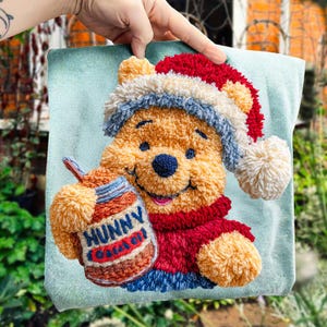 Puede incluir: Un cojín cuadrado azul claro con un diseño texturizado de Winnie the Pooh. Pooh lleva un gorro de Papá Noel rojo y blanco y sostiene un tarro de "Hunny". El diseño está hecho con hilo texturizado en tonos amarillos, rojos, azules y blancos.