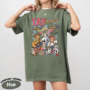 May include: A moss green short-sleeve t-shirt featuring a graphic print with the text TAYstory I KNEW IT I KNEW YOU Taylor. The design includes three illustrated figures in various outfits, including a space suit and cowboy attire.