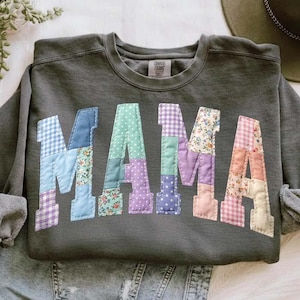 Op de afbeelding: Donkergrijze sweatshirt met het woord "MAMA" op de voorkant. De letters zijn gemaakt van patchworkstof in verschillende patronen en kleuren, waaronder blauw ruitjes, bloemenprints en stippen. De sweatshirt heeft lange mouwen en een ronde hals.