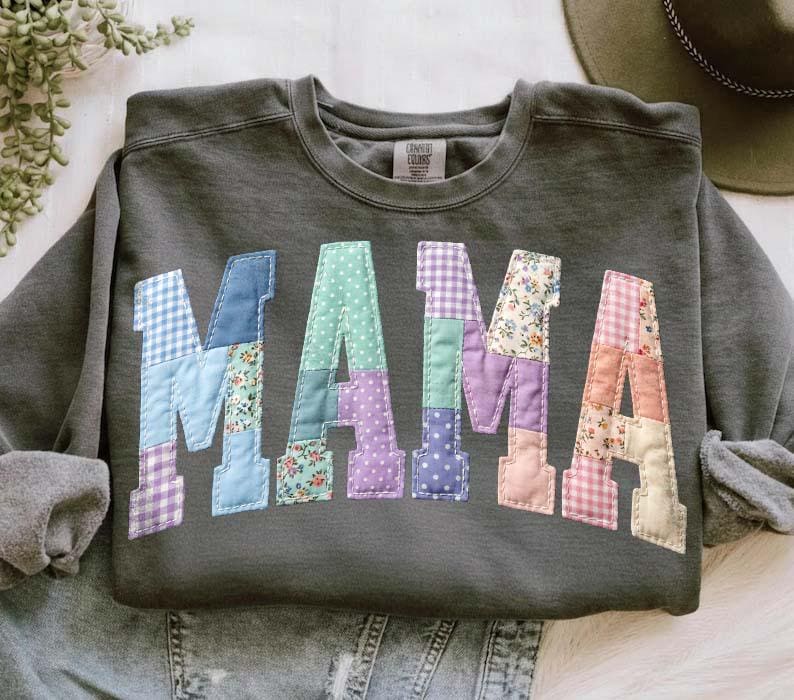 Op de afbeelding: Donkergrijze sweatshirt met het woord "MAMA" op de voorkant. De letters zijn gemaakt van patchworkstof in verschillende patronen en kleuren, waaronder blauw ruitjes, bloemenprints en stippen. De sweatshirt heeft lange mouwen en een ronde hals.