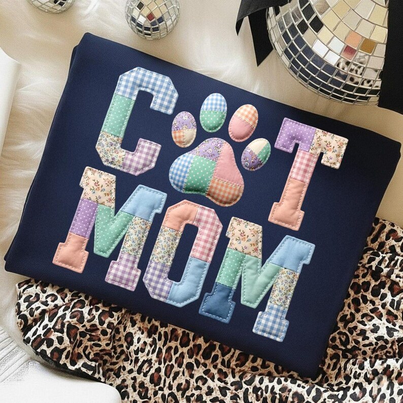 Pode incluir: Camisola azul marinho com as palavras "CAT MOM" em letras de patchwork e uma pegada de pata de patchwork. As letras e a pegada da pata s&atilde;o feitas de v&aacute;rios tecidos estampados, incluindo desenhos florais e xadrez. A camisola est&aacute; dobrada sobre uma superf&iacute;cie branca.