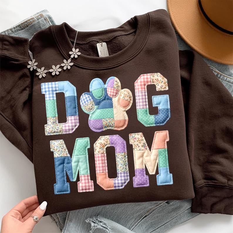以下が含まれることがあります： カラフルなパッチワーク生地で「DOG MOM」の文字がアップリケされた茶色のスウェットシャツ。「DOG」の「O」は足跡です。花型のペンダントが付いたシルバーのネックレスと茶色の帽子も写っています。