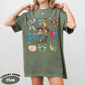 Op de afbeelding: Een olijfgroen t-shirt met korte mouwen en een grafische print van diverse stripfiguren, waaronder een space ranger, een cowgirl, een cowboy, een varken, een dinosaurus en een hond, afgewisseld met kleurrijke strikken, sterren en planeten.