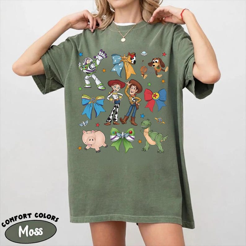 Op de afbeelding: Een olijfgroen t-shirt met korte mouwen en een grafische print van diverse stripfiguren, waaronder een space ranger, een cowgirl, een cowboy, een varken, een dinosaurus en een hond, afgewisseld met kleurrijke strikken, sterren en planeten.