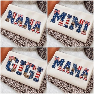 May include: A white fabric item featuring appliqued letters spelling NANA, MIMI, GIGI, or MAMA. The letters are crafted from blue fabric with white stars, red and white stripes, and leopard print patterns, accented with small red and white striped bows.