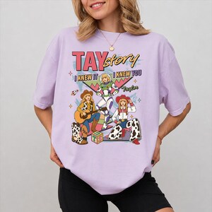 May include: A light purple t-shirt featuring a graphic print with the text TAYstory, I KNEW IT, I KNEW YOU, and Taylor. The design includes three illustrated figures in cowboy and space-themed outfits, a guitar, a rocket, and a block with the letters TS.
