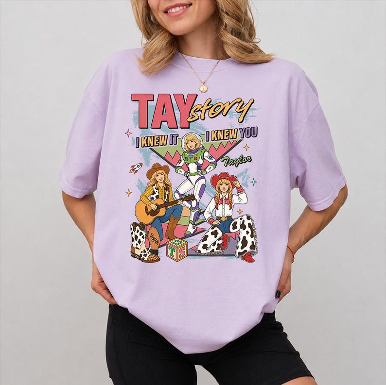 May include: A light purple t-shirt featuring a graphic print with the text TAYstory, I KNEW IT, I KNEW YOU, and Taylor. The design includes three illustrated figures in cowboy and space-themed outfits, a guitar, a rocket, and a block with the letters TS.