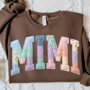 Op de afbeelding: Een bruine sweatshirt met het woord "MIMI" op de voorkant. De letters zijn gemaakt van patchworkstof in verschillende kleuren en patronen, waaronder bloemen, ruitjes en stippen. De sweatshirt heeft een ronde hals en lange mouwen.