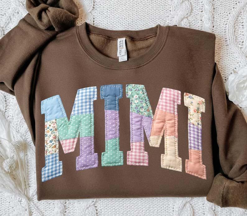 Op de afbeelding: Een bruine sweatshirt met het woord "MIMI" op de voorkant. De letters zijn gemaakt van patchworkstof in verschillende kleuren en patronen, waaronder bloemen, ruitjes en stippen. De sweatshirt heeft een ronde hals en lange mouwen.