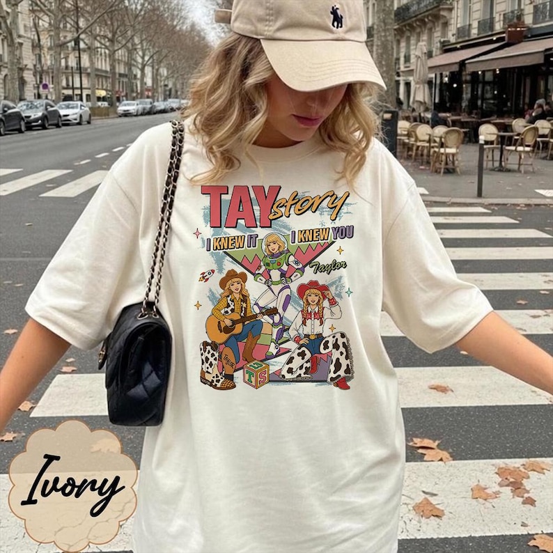 May include: An ivory t-shirt featuring a graphic print with the text TAYstory, I KNEW IT, I KNEW YOU, and Taylor. The design includes illustrations of three characters in western-style outfits, a guitar, a rocket, and a block with the letters TS.