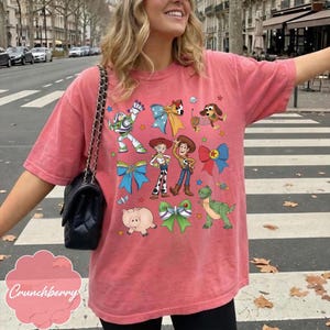 Op de afbeelding: Een koraalkleurig T-shirt met korte mouwen en een grafische print van diverse stripfiguren, waaronder een space ranger, een cowgirl, een cowboy, een varken, een dinosaurus en een hond, afgewisseld met kleurrijke strikken en ster- en planeeticonen.