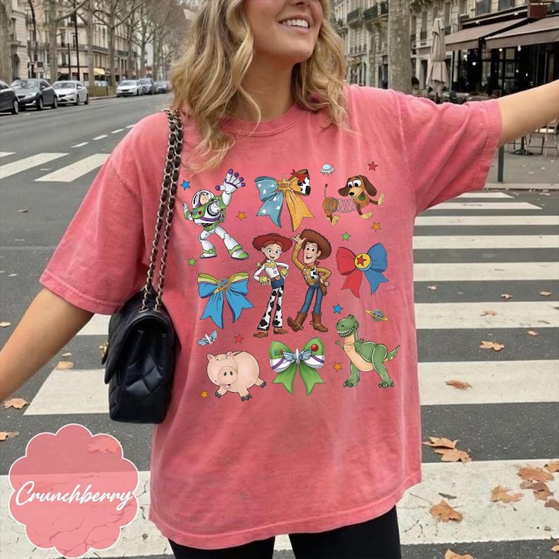 Op de afbeelding: Een koraalkleurig T-shirt met korte mouwen en een grafische print van diverse stripfiguren, waaronder een space ranger, een cowgirl, een cowboy, een varken, een dinosaurus en een hond, afgewisseld met kleurrijke strikken en ster- en planeeticonen.