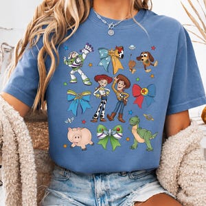 Op de afbeelding: Een blauw t-shirt met een grafische print van Toy Story-personages, waaronder Buzz Lightyear, Jessie, Woody, Slinky Dog, Rex en Hamm, afgewisseld met kleurrijke decoratieve strikken en kleine sterren verspreid over de voorkant.