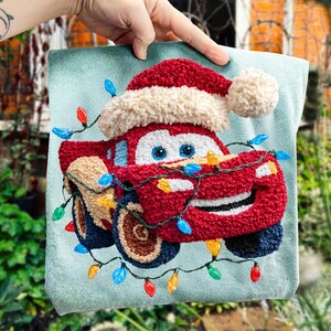 May include: A red car with a Santa hat and string lights on a light blue background. The car has blue eyes and a smiling face. The wheels are brown and blue. The lights are red, blue, yellow, and green.
