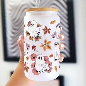 Könnte beinhalten: Ein weißer Becher mit einem Holzdeckel und einem Strohhalm. Der Becher hat ein Muster aus Geistern, Blumen und Blättern in Rosa-, Orange- und Brauntönen.
