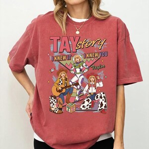 May include: A brick red short-sleeve t-shirt featuring a graphic print with the text TAYstory, I KNEW IT, I KNEW YOU, and Taylor. The illustration depicts three figures in various outfits, including a space suit and western attire with cow print.