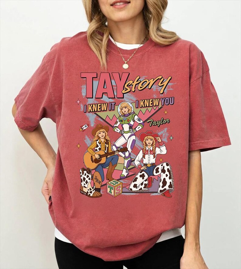 May include: A brick red short-sleeve t-shirt featuring a graphic print with the text TAYstory, I KNEW IT, I KNEW YOU, and Taylor. The illustration depicts three figures in various outfits, including a space suit and western attire with cow print.