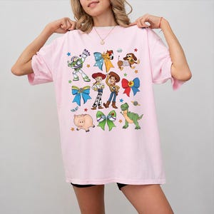 Op de afbeelding: Een lichtroze T-shirt met korte mouwen en een kleurrijke grafische print van diverse geanimeerde personages, waaronder een space ranger, een cowgirl, een cowboy, een varken, een dinosaurus en een hond, afgewisseld met strikken en kleine sterren.