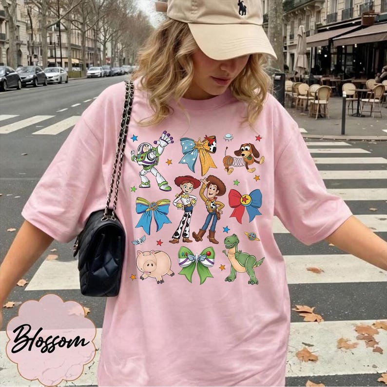 Op de afbeelding: Een lichtroze t-shirt met een grafische print van diverse tekenfilmfiguren, waaronder een space ranger, een cowgirl, een cowboy, een hond, een varken en een dinosaurus, afgewisseld met kleurrijke strikken en kleine sterretjes.