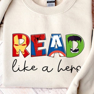 May include: A beige sweatshirt with the word "READ" in large, colorful letters. Each letter features a different superhero character: Iron Man, Spiderman, Captain America, and Hulk. The text "like a hero" is written in black below the word "READ".