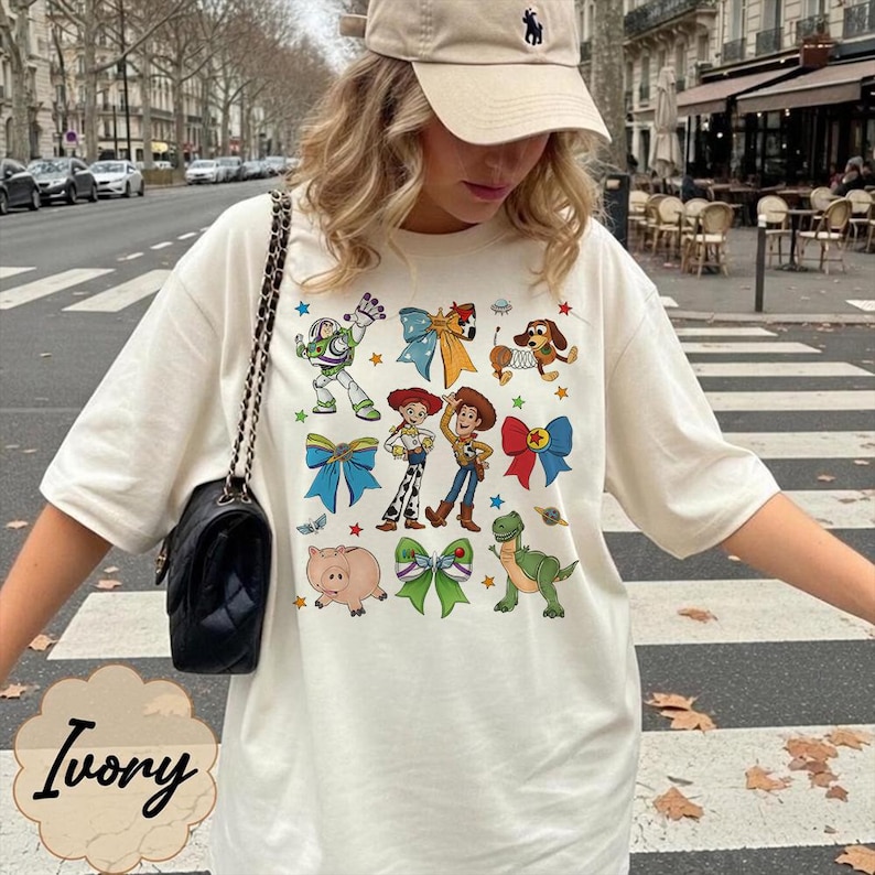 Op de afbeelding: Een ivoorkleurig t-shirt met een grafische print van diverse stripfiguren, waaronder een space ranger, een cowboy, een cowgirl, een dinosaurus, een varken en een hond, afgewisseld met kleurrijke strikken en sterren.