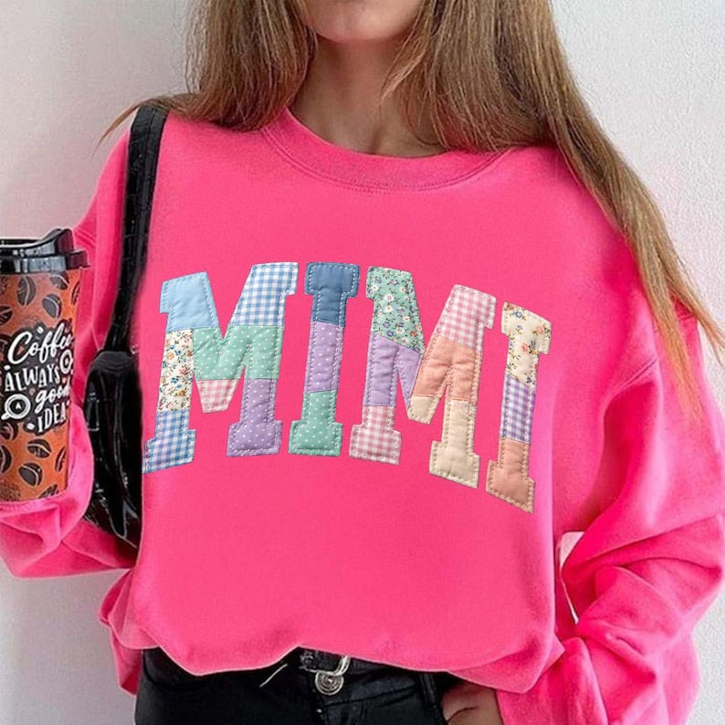Op de afbeelding: Een felroze sweatshirt met het woord "MIMI" in een patchwork-ontwerp op de voorkant. De letters zijn in verschillende pastelkleuren en patronen. Een zwarte schoudertas en een koffiekopje met tekst zijn ook zichtbaar.