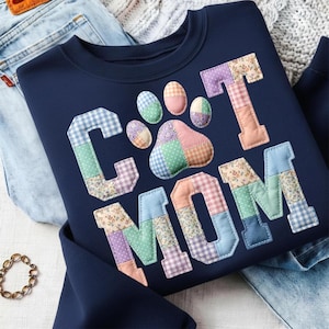 May include: Navy blue sweatshirt with the words "CAT MOM" in patchwork letters and a paw print design. The letters and paw print are made of various patterned fabrics in pastel colors. The sweatshirt is folded on top of a pair of blue jeans.