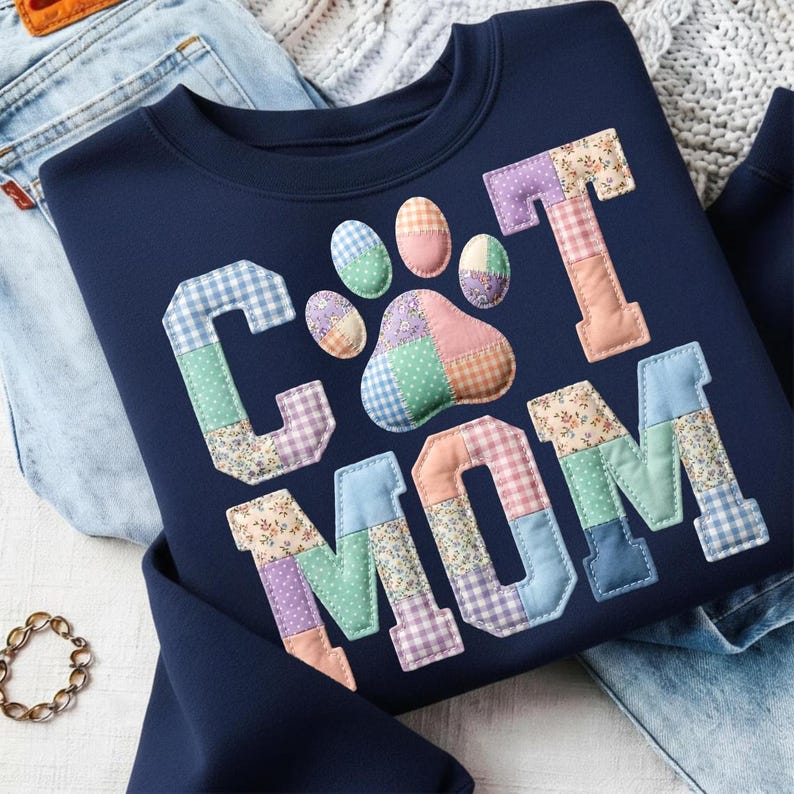 Pode incluir: Camisola azul marinho com as palavras "CAT MOM" em letras de patchwork e um desenho de pegada. As letras e a pegada s&atilde;o feitas de v&aacute;rios tecidos estampados em cores pastel. A camisola est&aacute; dobrada em cima de um par de jeans.