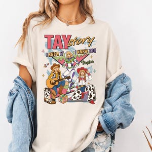 May include: A cream-colored t-shirt featuring a graphic print with the text TAYstory, I KNEW IT, and I KNEW YOU. The design includes three illustrated figures, a space ranger, and two cowgirls, along with a guitar, a toy block, and stars.