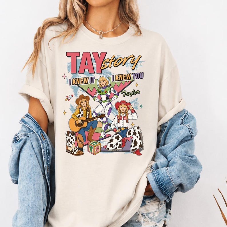 May include: A cream-colored t-shirt featuring a graphic print with the text TAYstory, I KNEW IT, and I KNEW YOU. The design includes three illustrated figures, a space ranger, and two cowgirls, along with a guitar, a toy block, and stars.