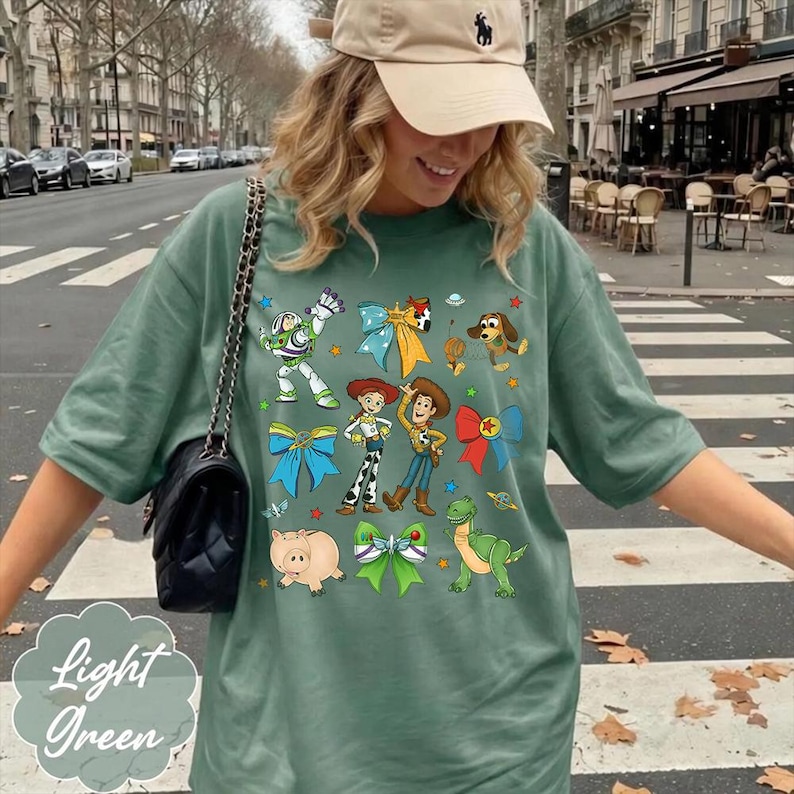 Op de afbeelding: Een saliegroen T-shirt met korte mouwen voorzien van een grafische print van diverse Toy Story-personages, waaronder Buzz Lightyear, Woody, Jessie, Rex, Slinky Dog en Hamm, afgewisseld met kleurrijke decoratieve strikken en sterren.