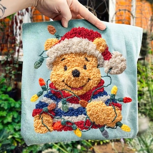 Puede incluir: Un cojín cuadrado azul claro con un diseño de Winnie the Pooh. El oso lleva un gorro de Papá Noel y un suéter a rayas, y está envuelto en luces navideñas. El diseño está hecho con bordado texturizado.