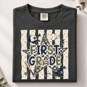 以下が含まれることがあります： FIRST GRADEという文字がプリントされたチャコールグレーのTシャツ。大きな星、野球帽、野球ボール、木製バット、アメリカンフットボールのヘルメットが、縦縞とドットの背景に描かれたグラフィックデザインが特徴です。