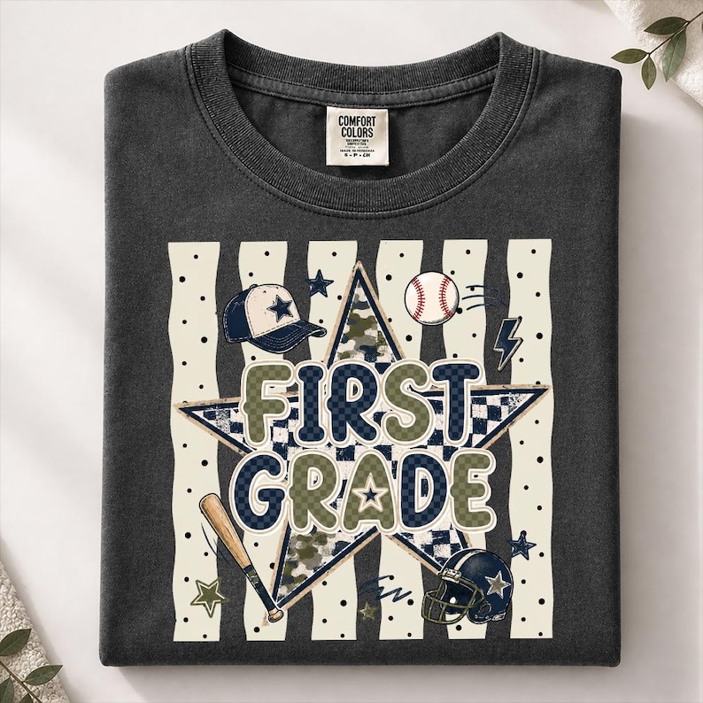 以下が含まれることがあります： FIRST GRADEという文字がプリントされたチャコールグレーのTシャツ。大きな星、野球帽、野球ボール、木製バット、アメリカンフットボールのヘルメットが、縦縞とドットの背景に描かれたグラフィックデザインが特徴です。