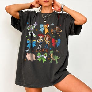 Op de afbeelding: Een antracietgrijs T-shirt met korte mouwen en een grafische print van diverse geanimeerde personages, waaronder een space ranger, een cowgirl, een cowboy, een varken, een dinosaurus en een hond, afgewisseld met kleurrijke strikken en sterren.
