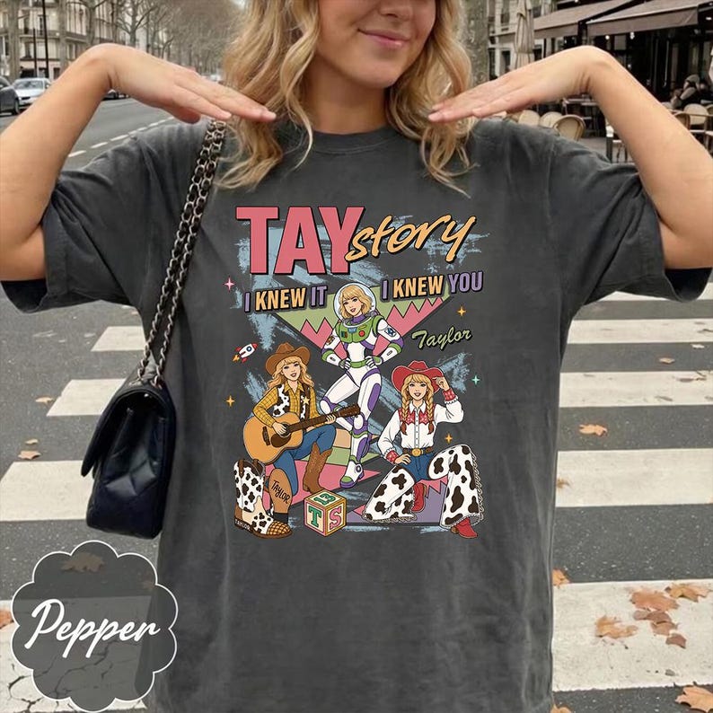 May include: A charcoal gray short-sleeve t-shirt featuring a graphic print with the text TAYstory I KNEW IT I KNEW YOU Taylor. The design includes three illustrated characters, a guitar, a toy block with TS, and a rocket ship in vibrant colors.
