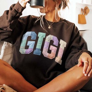 Op de afbeelding: Een donkerbruine sweatshirt met de naam "GIGI" in grote letters in patchwork-stijl op de voorkant. De letters zijn gemaakt van verschillende stoffen met patronen in tinten blauw, groen, paars en roze. De sweatshirt heeft lange mouwen.