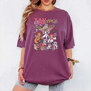 May include: A plum-colored t-shirt featuring a graphic print with the text TAY story I KNEW IT I KNEW YOU Taylor. The design includes three illustrated figures in cowboy and space-themed outfits, a guitar, and a block with the letters TS on it.