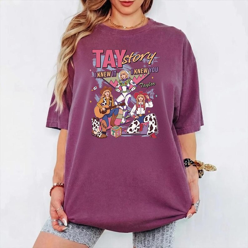 May include: A plum-colored t-shirt featuring a graphic print with the text TAY story I KNEW IT I KNEW YOU Taylor. The design includes three illustrated figures in cowboy and space-themed outfits, a guitar, and a block with the letters TS on it.