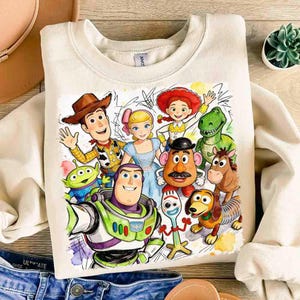 Pode incluir: Camisola creme com um gr&aacute;fico colorido de personagens do filme de anima&ccedil;&atilde;o Toy Story. O design inclui Woody, Buzz Lightyear, Jessie e outros personagens. A camisola tem gola redonda e mangas compridas.