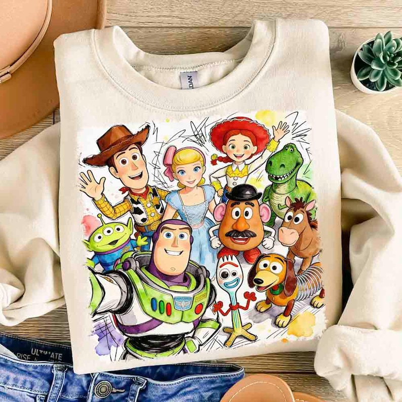 Pode incluir: Camisola creme com um gr&aacute;fico colorido de personagens do filme de anima&ccedil;&atilde;o Toy Story. O design inclui Woody, Buzz Lightyear, Jessie e outros personagens. A camisola tem gola redonda e mangas compridas.