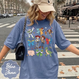 Op de afbeelding: Een blauw t-shirt met een grafische print van diverse stripfiguren, waaronder een space ranger, een cowgirl, een cowboy, een varken, een dinosaurus en een hond, afgewisseld met kleurrijke decoratieve strikken en kleine sterren en planeten.