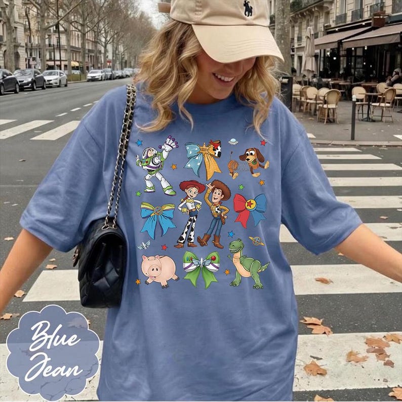 Op de afbeelding: Een blauw t-shirt met een grafische print van diverse stripfiguren, waaronder een space ranger, een cowgirl, een cowboy, een varken, een dinosaurus en een hond, afgewisseld met kleurrijke decoratieve strikken en kleine sterren en planeten.