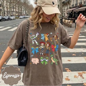Op de afbeelding: Een bruin t-shirt met korte mouwen en een grafische print van diverse stripfiguren, waaronder een space ranger, een cowboy, een cowgirl, een dinosaurus en een varken, afgewisseld met kleurrijke strikken en kleine ruimtevaart-iconen.