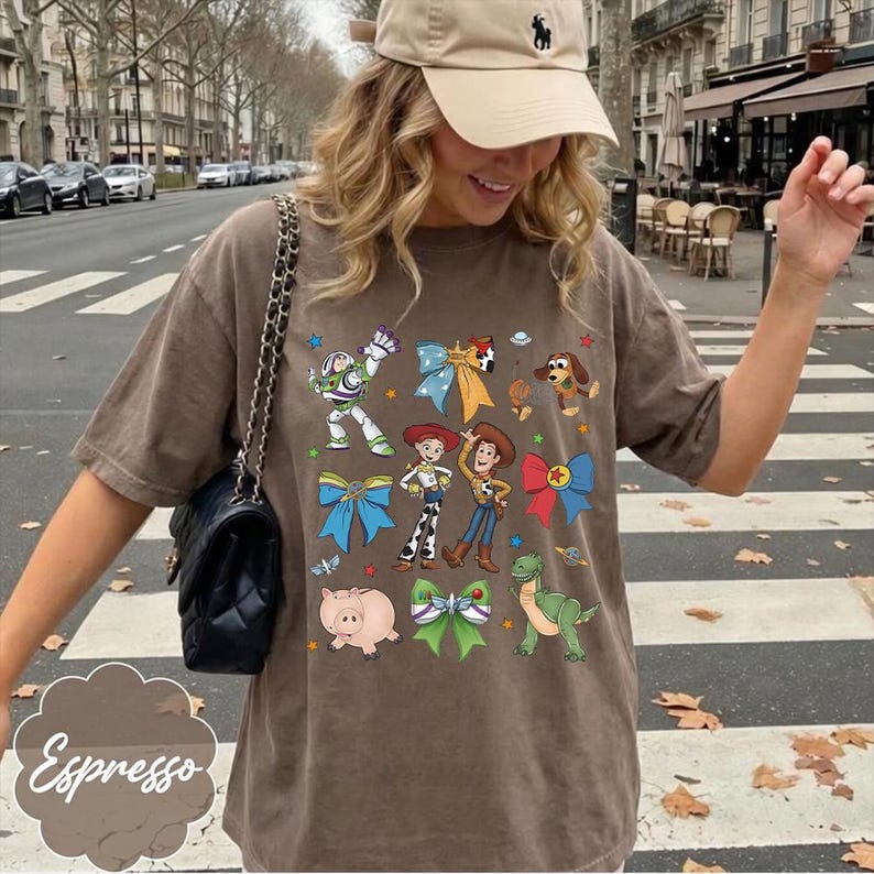 Op de afbeelding: Een bruin t-shirt met korte mouwen en een grafische print van diverse stripfiguren, waaronder een space ranger, een cowboy, een cowgirl, een dinosaurus en een varken, afgewisseld met kleurrijke strikken en kleine ruimtevaart-iconen.